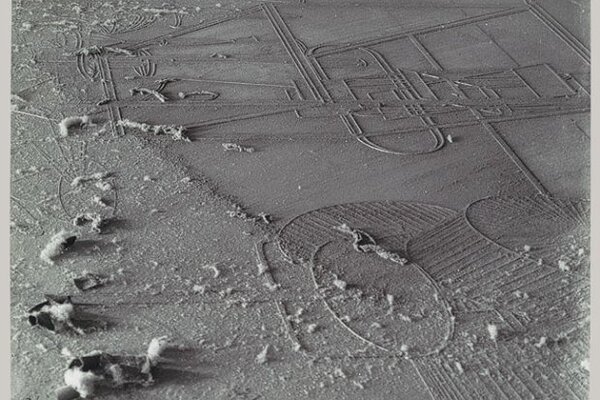 Schwarzweißfotografie einer staubbedeckten Glasplatte aus der Vogelperspektive. Die Oberfläche ist von einer feinen, ungleichmäßigen Staubschicht überzogen, die wie eine karge, mondähnliche Landschaft wirkt. In den Staub zeichnen sich geometrische Linien und Formen ab – gerade Strecken, Kreise und ovale Umrisse –, die technisch und konstruiert erscheinen. Die Linien wirken wie freigelegte Konturen unter der Staubschicht. Im linken unteren Bereich liegen mehrere kleine, weiche Staubballen oder Flocken, die sich von der flachen Fläche plastisch abheben. Rechts unten ist ein großer ovaler Bereich mit parallelen, gerillten Strukturen sichtbar, die an Felder oder technische Schraffuren erinnern. Am unteren Bildrand befindet sich eine schmale weiße Rahmung mit handschriftlichen Notizen und einer Signatur. 