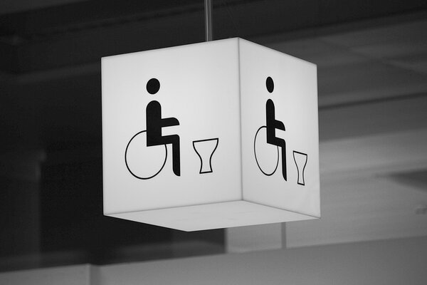 Schwarz-weiß-Fotografie von Leuchtkubus mit Piktogramm-Kombination. Diese zeigt eine Person in Rollstuhl neben dem Umriss einer Toilette. Der Kubus ist an einer Zimmerdecke angebracht.