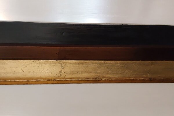 Das Foto "Gold, Braun, Schwarz" besteht aus drei senkrechten Streifen in Gold, Braun und Schwarz vor einem hellen Hintergrund. Die Farbe ist auf Holz gestrichen, dessen Struktur zu erkennen ist.