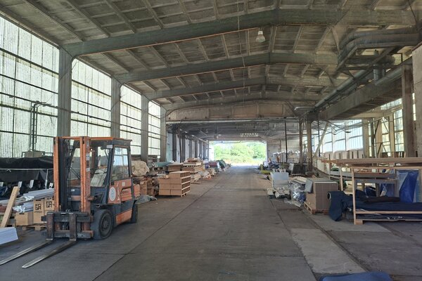 Blick in ein Materiallager einer Industriehalle. Links im Vordergrund steht ein orangefarbener Gabelstapler. Entlang beider lichtdurchlässigen Hallenseiten lagern unterschiedliche Materialien, Bauteile, Paletten und textile Elemente in offenen Regalen oder gestapelt auf dem Boden. Die Mitte der Halle bleibt als breite Fahr- und Bewegungsfläche frei und führt den Blick zu einer geöffneten Hallenseite im Hintergrund. 